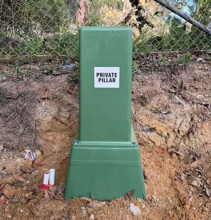 Green private supply pillar labelled PRIVATE PILLAR, set into a cut bank beside a fence line